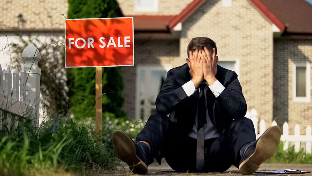 Frustrated Real Estate agent sitting on ground outside of a home for sale